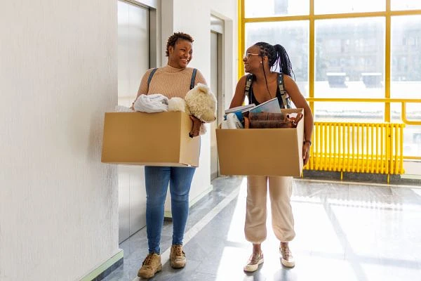 Two people are smiling and carrying boxes while walking down a hallway.