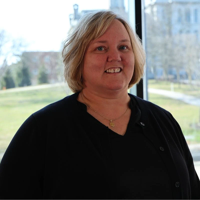 A person is smiling indoors with a scenic view of a park-like setting in the background.